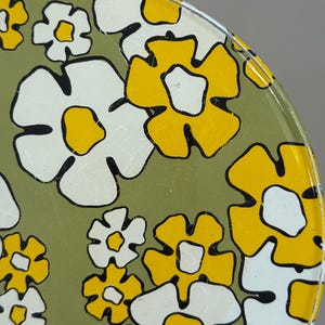 May include: Close-up of a vintage metal tray with a floral pattern. The design features large white and yellow flowers with black outlines on a green background. The tray has a slightly distressed appearance.