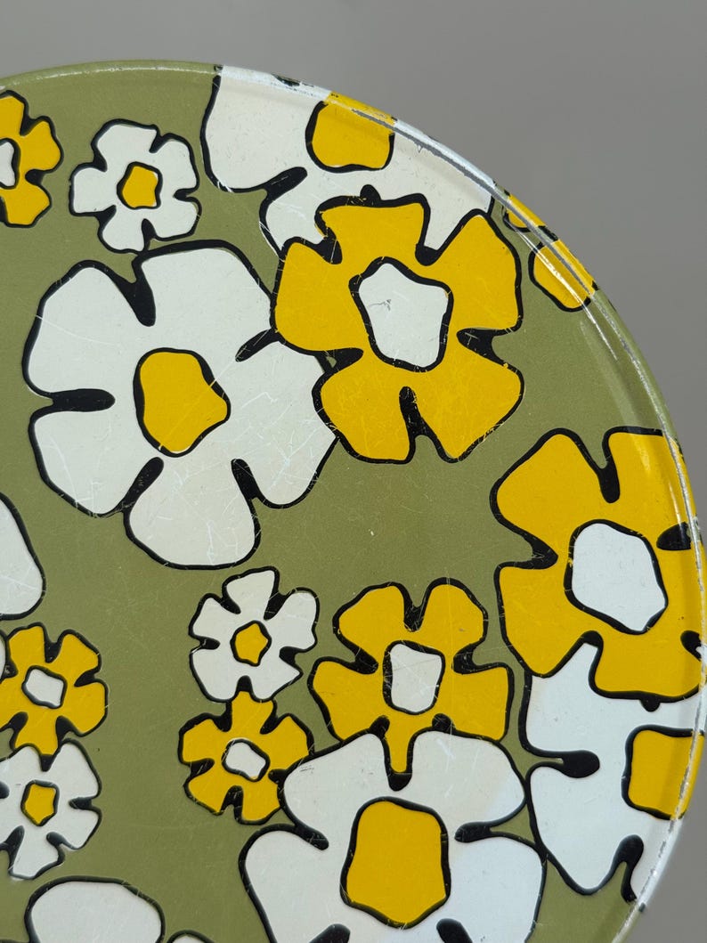May include: Close-up of a vintage metal tray with a floral pattern. The design features large white and yellow flowers with black outlines on a green background. The tray has a slightly distressed appearance.