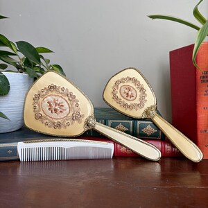 May include: A vanity set consisting of two cream-coloured hand mirrors and a white plastic comb. Each mirror features an oval centre panel with embroidered floral details, surrounded by a decorative gold-tone metal frame and a matching handle.