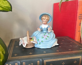 May include: A porcelain figurine of a girl in a blue dress and hat holding a basket with fruit. The figurine is sitting on a stack of books with the title "Little Women" visible.