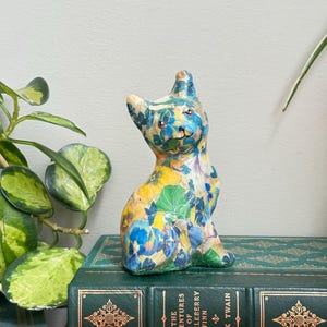 Pode incluir: Uma pequena estatueta de gato coberta com um padrão floral colorido com pétalas amarelas, azuis e verdes. O gato está sentado na vertical com orelhas pontudas e um rosto pintado, posicionado sobre um livro verde intitulado The Adventures of Huckleberry Finn.
