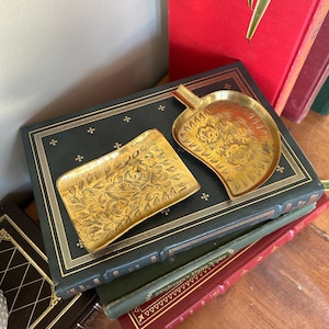 May include: Two small, gold-colored metal trays with intricate floral designs. The trays are sitting on top of a stack of books.