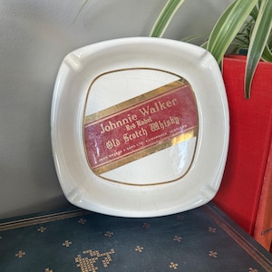 Op de afbeelding: Witte keramische asbak met een Johnnie Walker Red Label Old Scotch Whisky label. Het label is rood met gouden letters en een gouden rand.