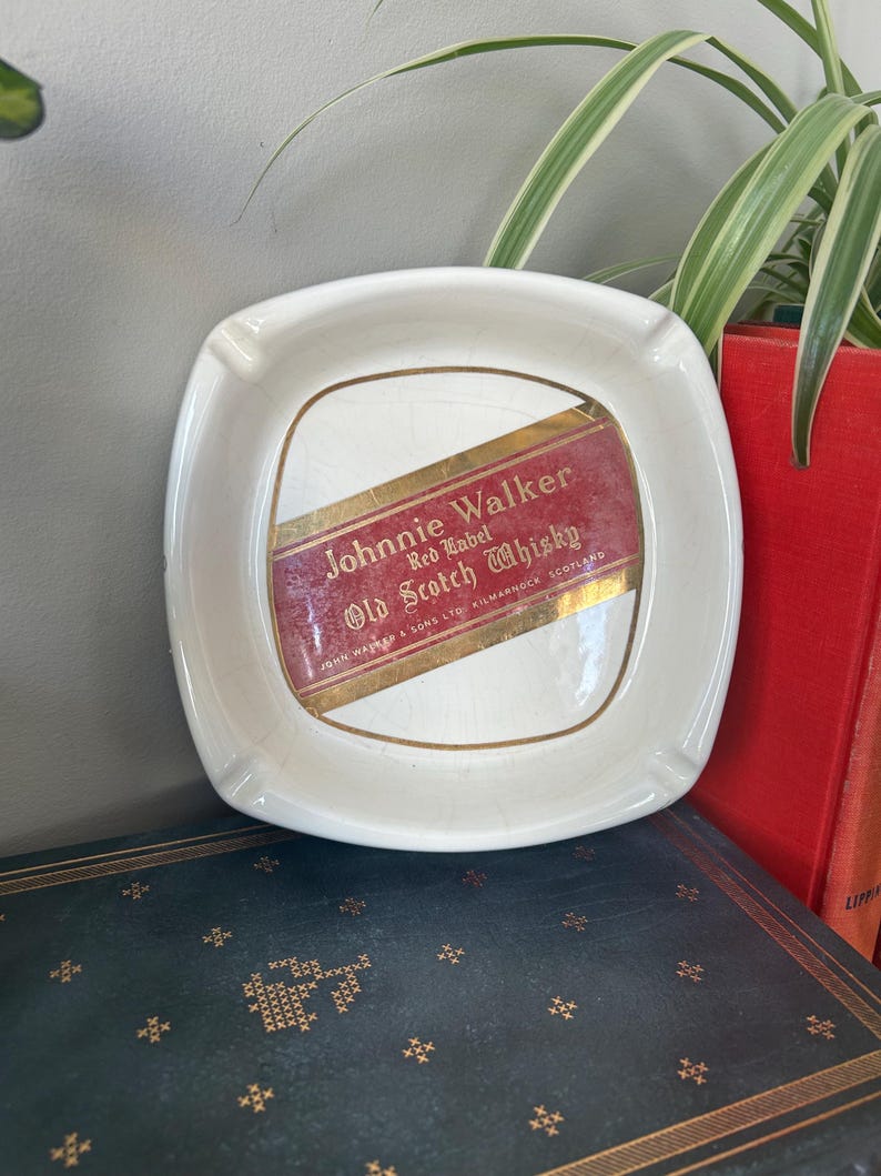 Op de afbeelding: Witte keramische asbak met een Johnnie Walker Red Label Old Scotch Whisky label. Het label is rood met gouden letters en een gouden rand.