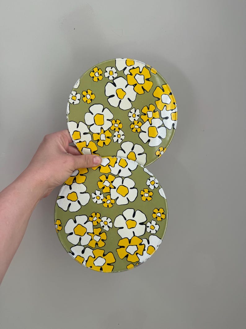 May include: Two round, floral-patterned plates. The plates feature a green background with white and yellow daisy-like flowers. The plates are held up against a gray background.
