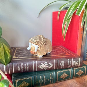 May include: A miniature tan musk ox figure with a textured coat and white horns sits atop a stack of classic books. The books are brown and green with gold lettering. A red book and green plants are in the background.