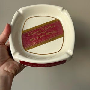 Op de afbeelding: Witte keramische asbak met een rood en gouden Johnnie Walker Red Label Old Scotch Whisky label. De asbak heeft een Johnnie Walker logo op de onderkant.