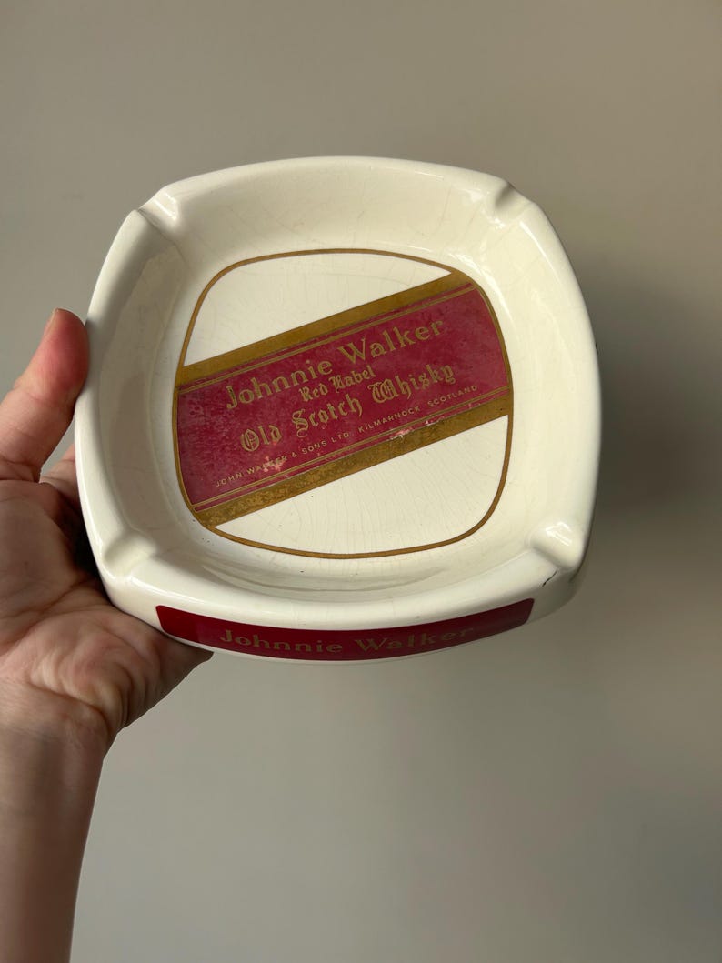 Op de afbeelding: Witte keramische asbak met een rood en gouden Johnnie Walker Red Label Old Scotch Whisky label. De asbak heeft een Johnnie Walker logo op de onderkant.