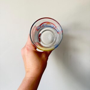 May include: Clear glass with a colorful design around the rim. The glass has red, blue, and yellow accents. The glass is empty and held up against a white background.