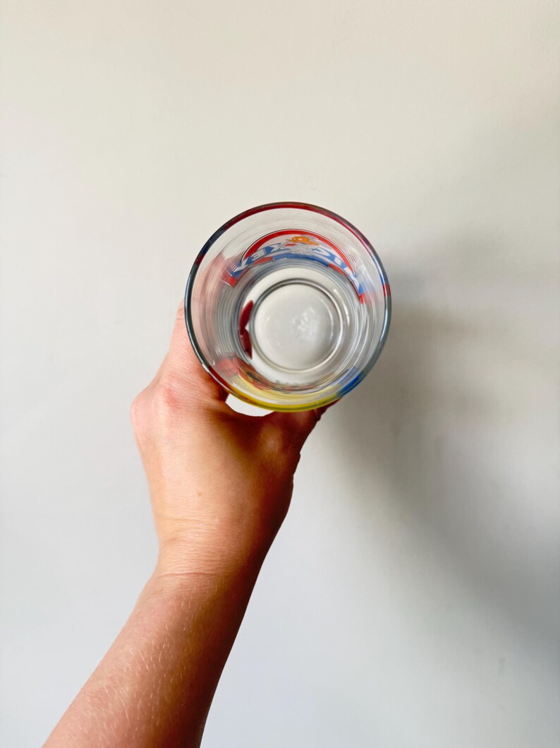May include: Clear glass with a colorful design around the rim. The glass has red, blue, and yellow accents. The glass is empty and held up against a white background.