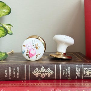 Op de afbeelding: Twee decoratieve porseleinen deurknoppen. Eén is rond met een bloemenontwerp in roze, blauw en goud. De andere is wit met een gouden basis. Beide liggen op een stapel antieke boeken met gouden letters.