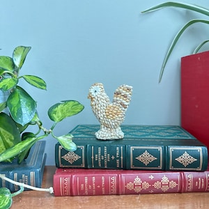May include: A small, textured figurine of a bird crafted from various small seashells in shades of cream, tan, and brown. The bird has a rounded body, a short tail, and a small eye, resting on a base made of clustered shells.