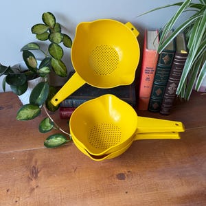 Puede incluir: Dos coladores de plástico amarillo brillante con asas apilados sobre una superficie de madera. Cada colador tiene un fondo perforado para escurrir líquidos. Estos coladores forman parte de un juego de utensilios de cocina.