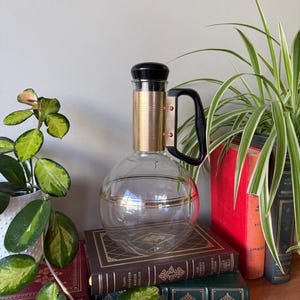 Peut inclure: Une carafe en verre transparent avec une bande dorée au milieu et un couvercle et une poignée noirs. La carafe est posée sur une pile de livres avec des accents dorés.
