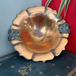 May include: A copper bowl with a scalloped edge and two ornate handles. The bowl has a shiny, slightly tarnished finish.