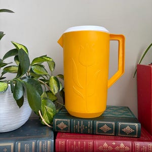 Puede incluir: Una jarra de plástico de color amarillo brillante con un diseño de tulipán moldeado en la parte frontal, una tapa blanca y un asa resistente. La jarra tiene un cuerpo cilíndrico con una base ligeramente cónica y un pico vertedor.