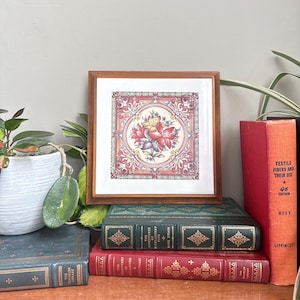May include: A framed floral tile with a brown wooden frame. The tile features a central flower design in red, pink, and yellow, surrounded by ornate patterns. The framed tile rests on a stack of vintage books with green and red covers.
