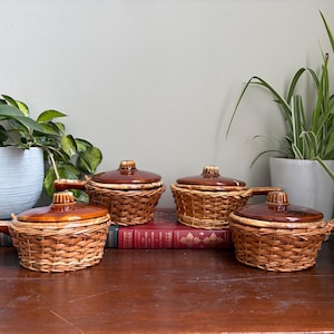 May include: Four round ceramic dishes with brown glazed lids and handles, each encased in a woven wicker basket holder. The dishes are arranged in a group, with two in the front and two slightly behind, resting on a dark wooden surface.