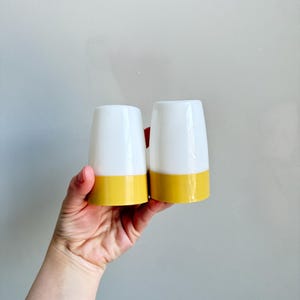 May include: Two white and yellow tumblers are held up against a gray background. The tumblers have a white upper section and a yellow base. The shape is slightly tapered, wider at the top and narrower at the bottom.