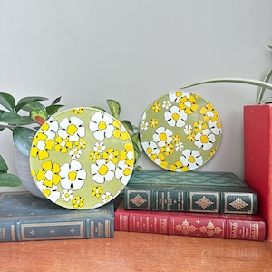 May include: Two circular coasters with a retro floral pattern. The coasters have a light green base with white and yellow flowers. They are displayed on a stack of vintage books with green and red covers.