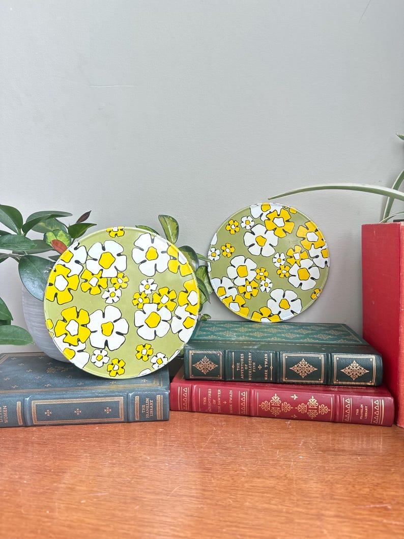 May include: Two round coasters with a retro floral design. The coasters feature a light green background with white and yellow flowers. They are placed on top of a stack of vintage books with green and red covers.