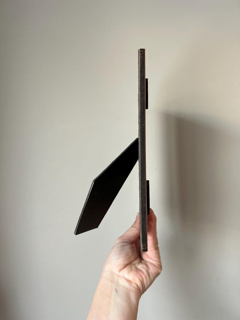 Puede incluir: Un marco de fotos de madera marr&oacute;n oscuro con un caballete negro. El marco est&aacute; de pie y lo sostiene una mano.