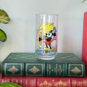 May include: A clear glass featuring Mickey Mouse at the helm of a ship, set against a yellow sun. Mickey is wearing a sailor hat, red shorts, and yellow shoes. The glass is on top of two antique books with green and red covers.
