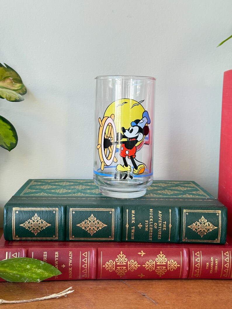 May include: A clear glass featuring Mickey Mouse at the helm of a ship, set against a yellow sun. Mickey is wearing a sailor hat, red shorts, and yellow shoes. The glass is on top of two antique books with green and red covers.