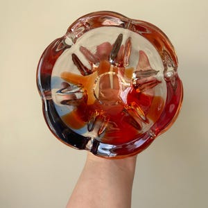 May include: A clear glass ashtray with a scalloped edge and a red, orange, and yellow swirl design.