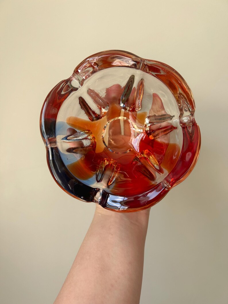 May include: A clear glass ashtray with a scalloped edge and a red, orange, and yellow swirl design.