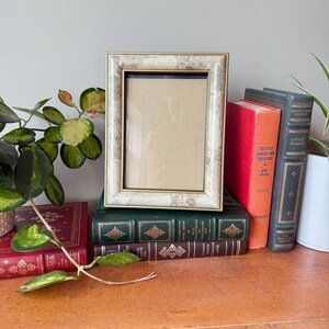Peut inclure: Un cadre photo rectangulaire avec un bord doré et un motif marbré. Il est posé sur une pile de livres anciens aux couvertures rouges, vertes et marron. Une plante verte à feuilles panachées est au premier plan.