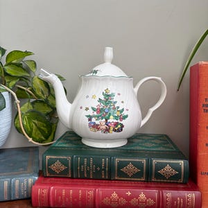 May include: A white ceramic teapot with a fluted body and a lid featuring a green trim. The front displays a colorful illustration of a decorated Christmas tree surrounded by wrapped gifts, stars, and ornaments, with a curved handle and spout.