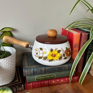 May include: White enamel saucepan with a brown lid and wooden handle. The saucepan features a floral design with yellow, white, and orange flowers. The saucepan is sitting on a stack of books.
