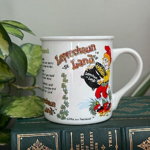 May include: A white ceramic mug featuring the text Leprechaun Land and an illustration of a leprechaun holding a black pot filled with gold coins. The mug includes a printed legend about leprechauns and the text Gifts from Ireland.