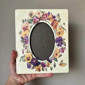 Puede incluir: Un marco de fotos ovalado blanco con un dise&ntilde;o floral con pensamientos morados, amarillos y verdes. El marco tiene un respaldo de cart&oacute;n marr&oacute;n.