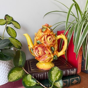 May include: A yellow and orange ceramic teapot with a floral design. The teapot is decorated with roses and other flowers. It is sitting on a stack of books.