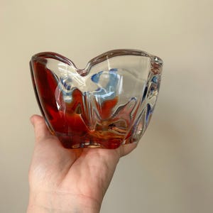 May include: A clear glass ashtray with a red and blue swirl design. The ashtray is shaped like a flower and has a wide, shallow bowl.