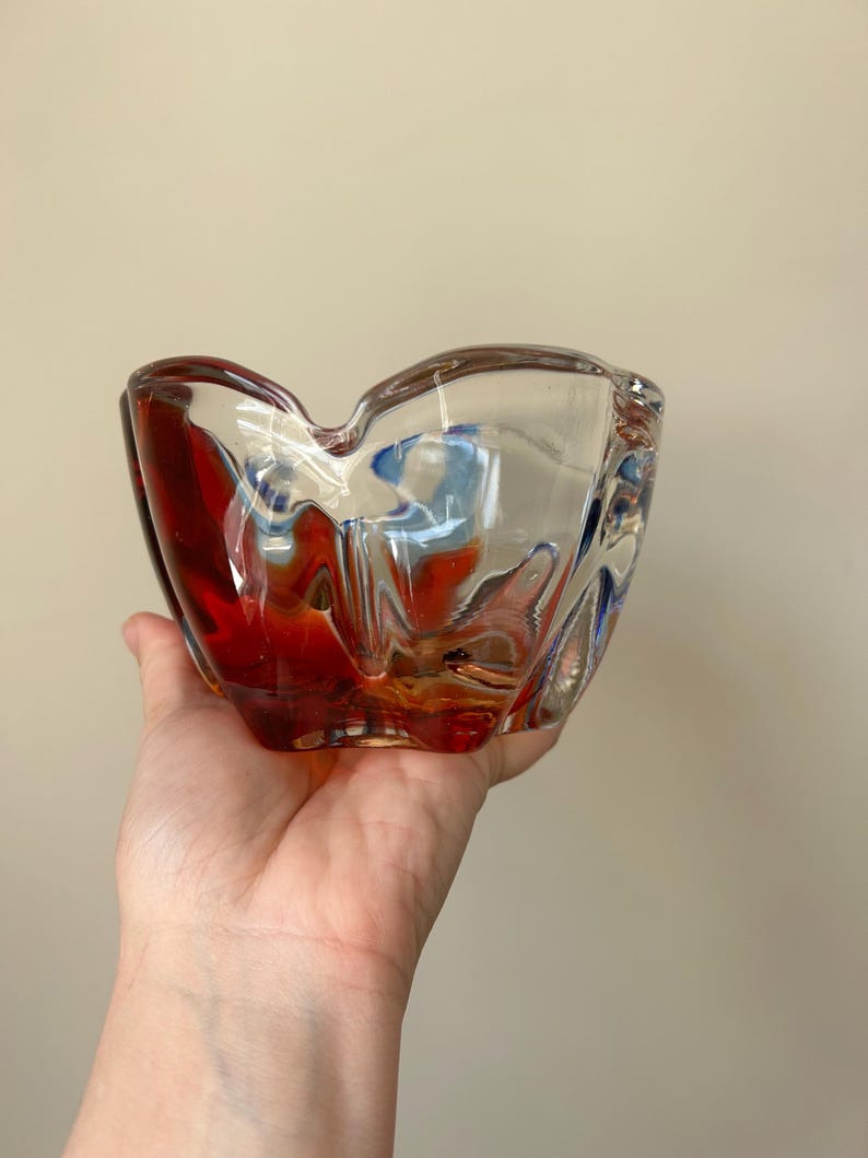 May include: A clear glass ashtray with a red and blue swirl design. The ashtray is shaped like a flower and has a wide, shallow bowl.
