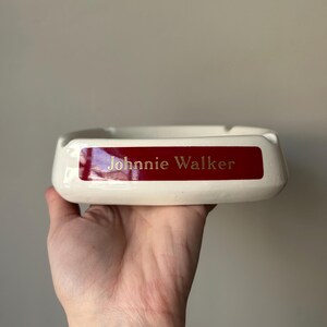 Op de afbeelding: Witte asbak van keramiek met een rode band en gouden opschrift "Johnnie Walker".