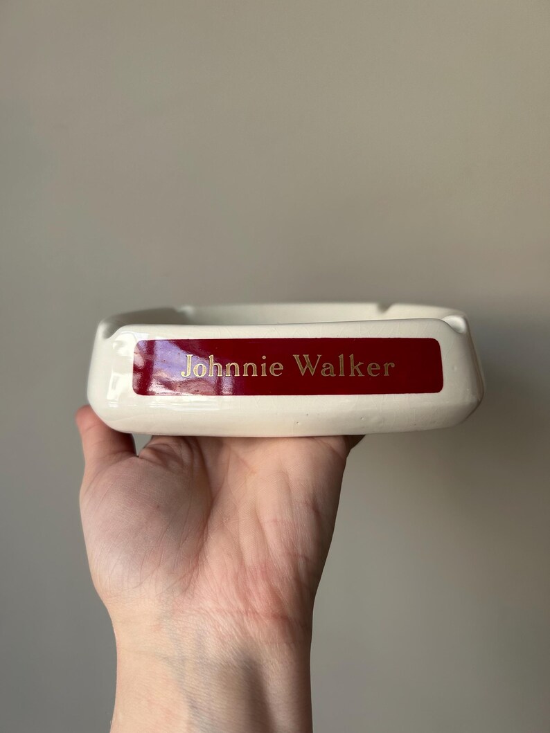 Op de afbeelding: Witte asbak van keramiek met een rode band en gouden opschrift "Johnnie Walker".