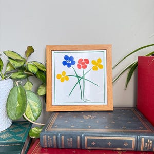 May include: A framed floral art print with blue, red, and yellow flowers on a white background. The print is in a light wood frame and is displayed on a stack of vintage books. The artwork is signed "Collins".