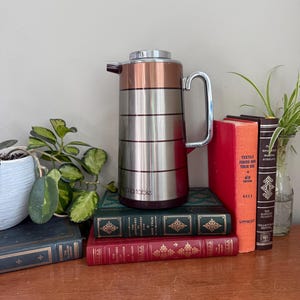 May include: A vintage stainless steel and copper carafe with a handle, resting on a stack of antique books. The carafe has the text "Rala table" on the side. Decorative plants are in the background.