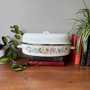 Pode incluir: Assadeira esmaltada branca com tampa, com um design floral em laranja, rosa e verde. A assadeira está em cima de uma pilha de livros antigos em uma superfície de madeira. Um item decorativo para cozinha.