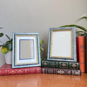 Könnte beinhalten: Zwei blaue Bilderrahmen mit goldenen Verzierungen und Seildetails. Die Rahmen werden auf einer Holzoberfläche mit einer Sammlung von Vintage-Büchern in verschiedenen Farben, darunter Rot, Grün und Braun, ausgestellt.