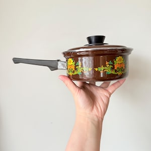 Op de afbeelding: Bruine steelpan met een zwarte handgreep en deksel. De steelpan is versierd met een bloemmotief in geel, oranje en groen. De handgreep is grijs en het deksel heeft een zwarte knop. Een vintage keukenartikel.