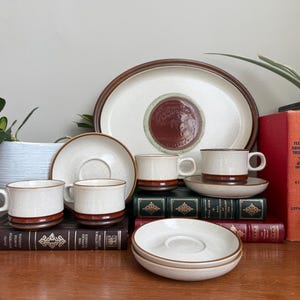 Puede incluir: Un conjunto de vajilla de cerámica vintage, que incluye tazas, platillos y una fuente para servir. Las piezas presentan un cuerpo color crema con una banda marrón en la base. Los artículos están dispuestos sobre una superficie de madera, con libros y una planta en maceta al fondo.