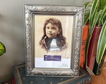 Può includere: Una cornice per foto argentata con un designo ornato e una bordura dorata. La cornice contiene una foto in bianco e nero di una bambina con i capelli ricci. La cornice è appoggiata su una pila di libri. La cornice è etichettata "CONNOISSEUR" e "5x7 in 13x18 cm".
