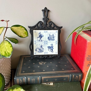 May include: A black cast iron trivet with a blue and white tile design featuring four scenes from a storybook. The tile is surrounded by a decorative black metal frame. The trivet is sitting on top of a stack of books.