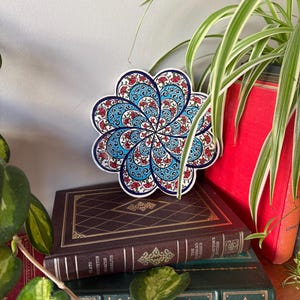 May include: A blue and red ceramic trivet with a floral design. The trivet is shaped like a flower and has a white border.