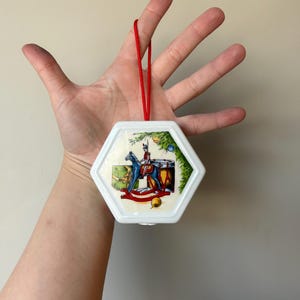 May include: White hexagonal Christmas bauble with a red string for hanging. The bauble features a colourful illustration of a toy soldier riding a rocking horse with a drum and a gift box.
